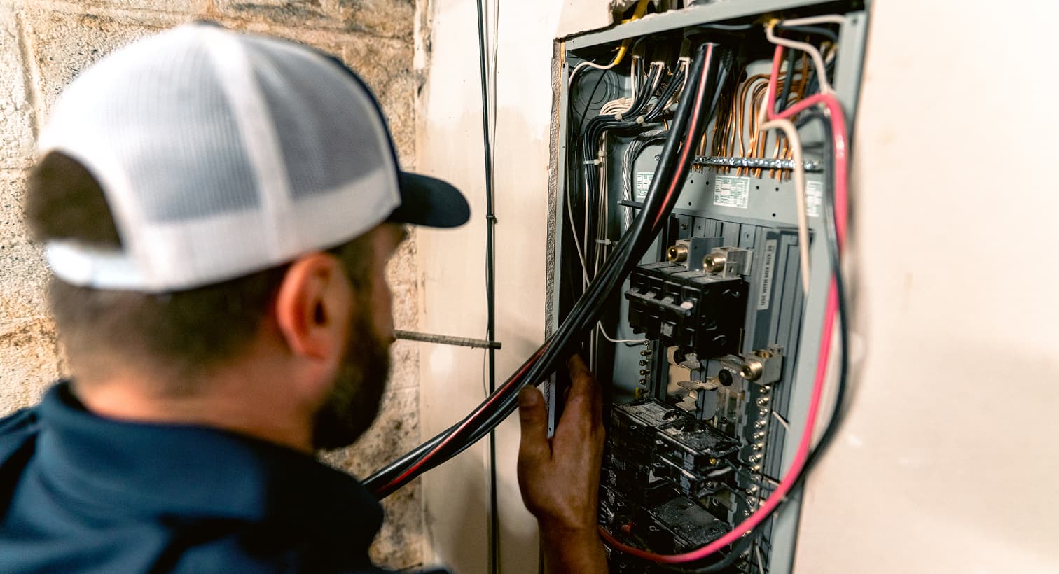 A professional electrician inspects and installs thick black and red electrical wires inside an open residential circuit breaker panel box on a basement wall.