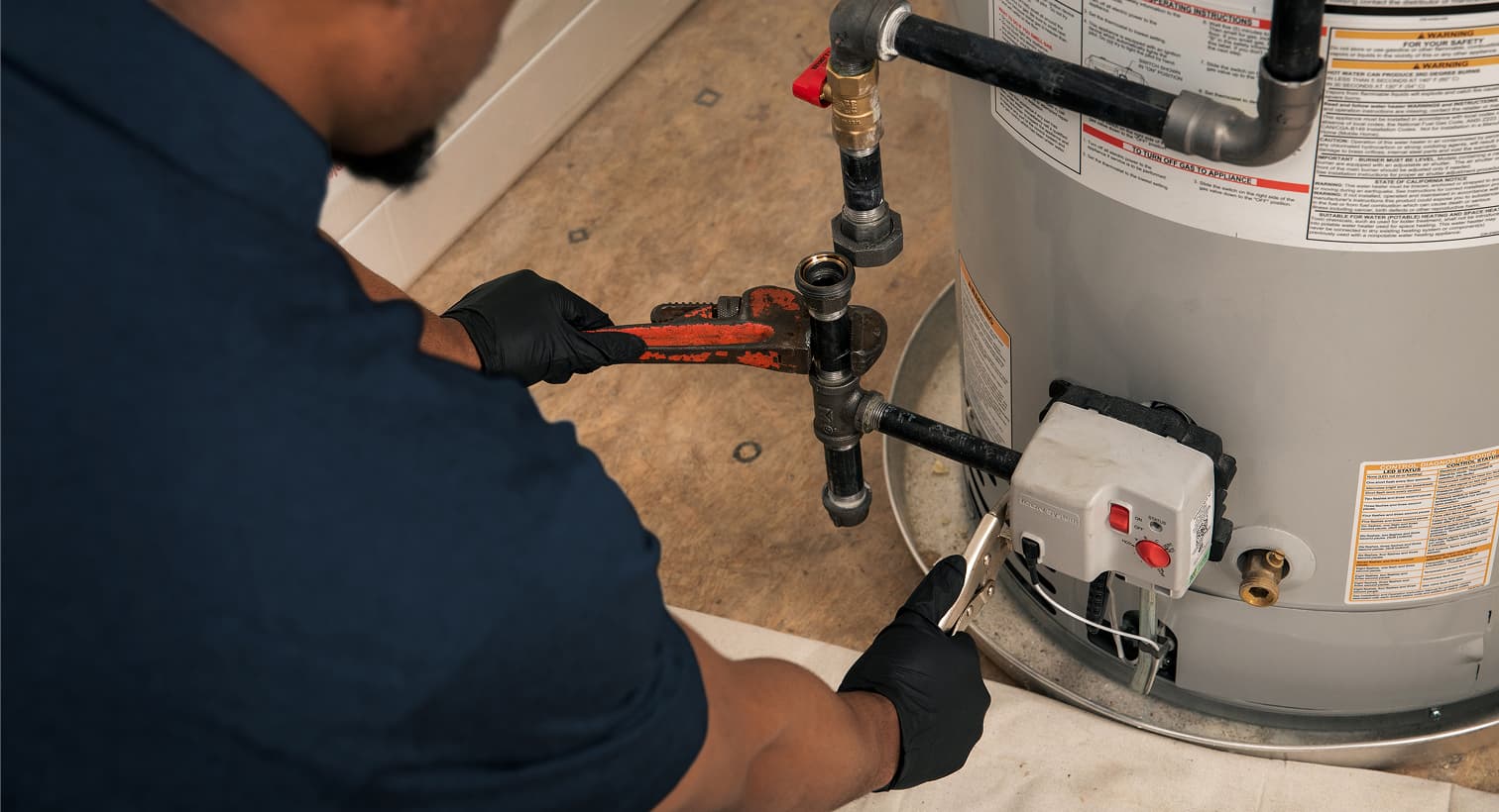 A Wm Henderson plumber wearing black gloves uses a pipe wrench to repair the gas line and control valve assembly on a residential water heater tank.